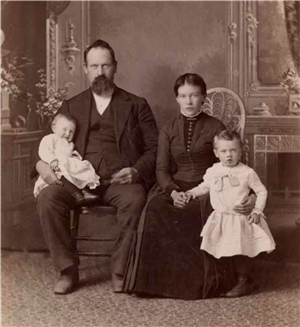 A white man and woman in suit and dress are seated holding a baby in arms and a child standing in front of the woman. They are seated in front of an ornate fireplace and mirror